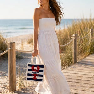 May include: A woman wearing a white strapless maxi dress and sunglasses walks on a wooden boardwalk. She carries a tote bag with navy blue and white stripes, a red lobster graphic, and the name "PERRY". The background shows a beach and ocean.