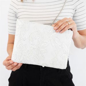 May include: White clutch with floral embroidery and a silver chain strap. The rectangular bag features a detailed, raised floral pattern across its surface. The clutch is held by a person wearing a striped shirt and black pants.