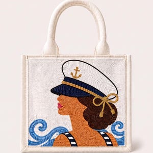 May include: A square off-white tote bag featuring a beaded design of a person wearing a white and navy blue captain's hat with a gold anchor emblem. The design includes brown hair, a striped top, and blue beaded waves on a white background.