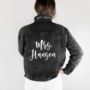 May include: A black denim jacket with white lettering that reads "Mrs. Hangen" on the back. The collar has "EST. 2027" printed in white. The jacket is a dark gray color.