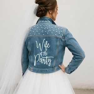 Könnte beinhalten: Hellblaue Jeansjacke mit Perlenbesatz auf den Schultern. Auf der Rückseite steht in weißer Schrift "Wife of the Party" und das Datum "10.12.2028". Die Jacke ist Teil eines Brautensembles, passend für eine Hochzeitsfeier.