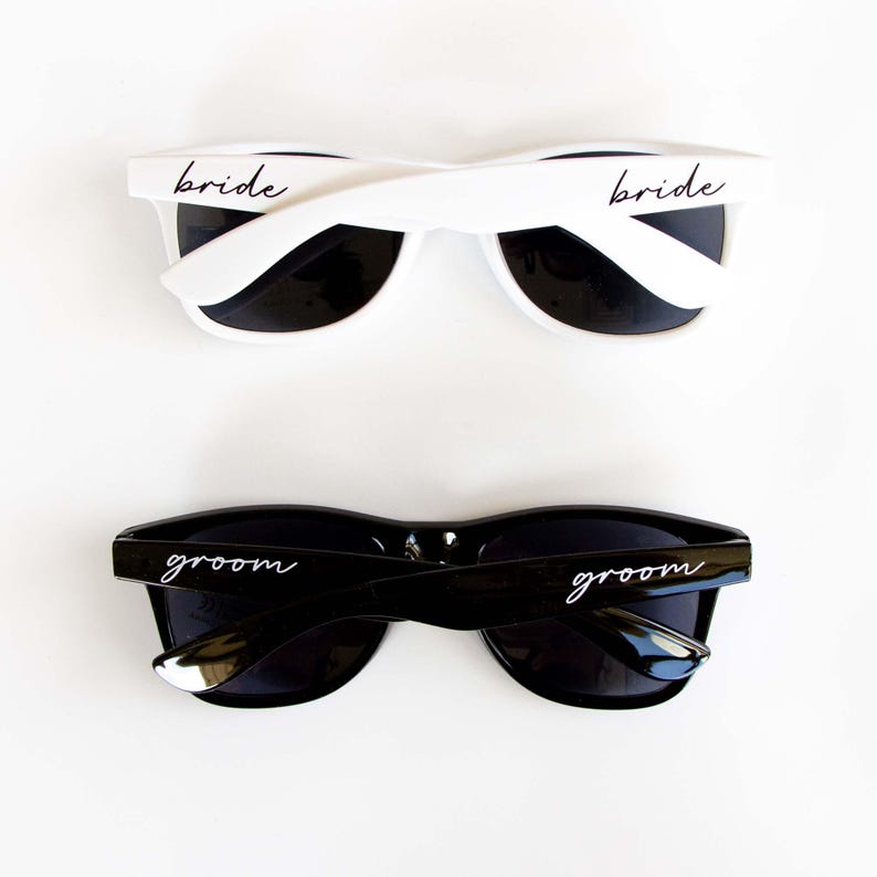 May include: Two pairs of sunglasses, one white with black lenses and the word "bride" on the frame, and one black with black lenses and the word "groom" on the frame.