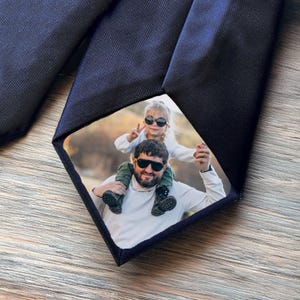 Puede incluir: Corbata azul marino con una foto personalizada de un hombre y un niño. La foto es un recuadro cuadrado en la corbata, que muestra al hombre llevando al niño sobre sus hombros. La corbata es de tela lisa y oscura.