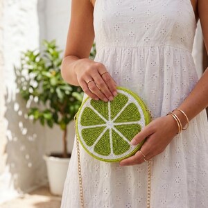 May include: A round, beaded crossbody bag shaped like a lime slice, featuring vibrant green and white glass beads arranged in a citrus wedge pattern, finished with a thin gold-tone metal chain strap for carrying over the shoulder.