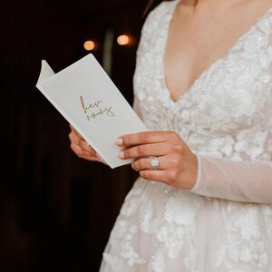 May include: A white notebook with the words "her vows" written in gold script. The notebook is being held by a person wearing a white lace dress and a diamond ring.