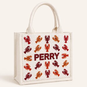 May include: Off-white tote bag with a beaded design of red, orange, and brown lobsters. The word "PERRY" is spelled out in red beads in the center of the bag. The bag has a white handle.