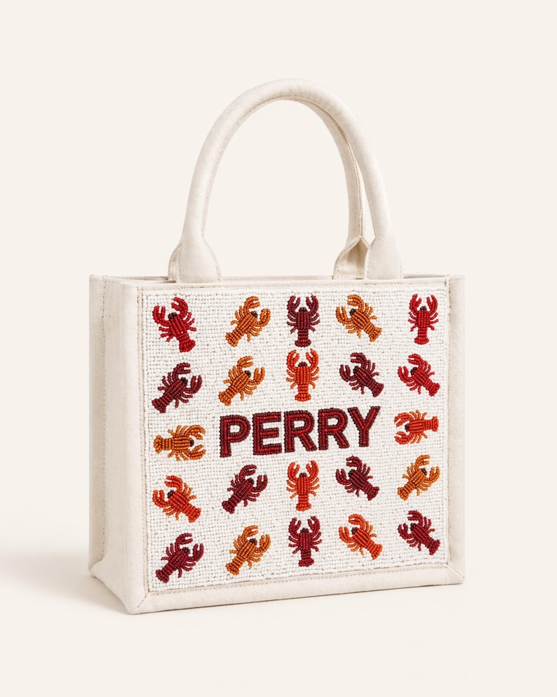 May include: Off-white tote bag with a beaded design of red, orange, and brown lobsters. The word "PERRY" is spelled out in red beads in the center of the bag. The bag has a white handle.