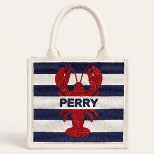May include: A small tote bag with a white handle and trim. The front features a navy and white striped pattern with a red lobster design and the name "PERRY" in white letters. The bag is likely made of fabric.