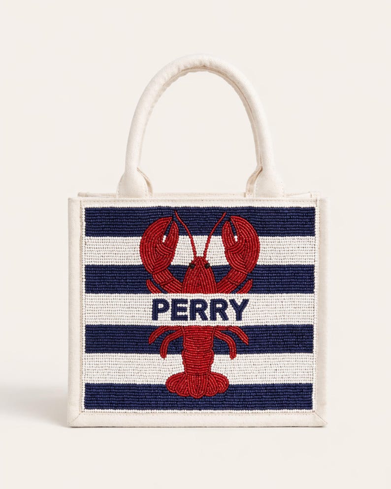 May include: A small tote bag with a white handle and trim. The front features a navy and white striped pattern with a red lobster design and the name "PERRY" in white letters. The bag is likely made of fabric.
