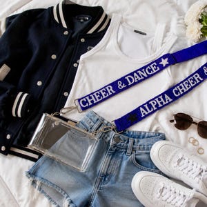 May include: A clear rectangular crossbody bag with a blue beaded strap featuring the text CHEER & DANCE and ALPHA CHEER. The bag is displayed with a navy blue varsity jacket, a white tank top, denim shorts, white trainers, and sunglasses.