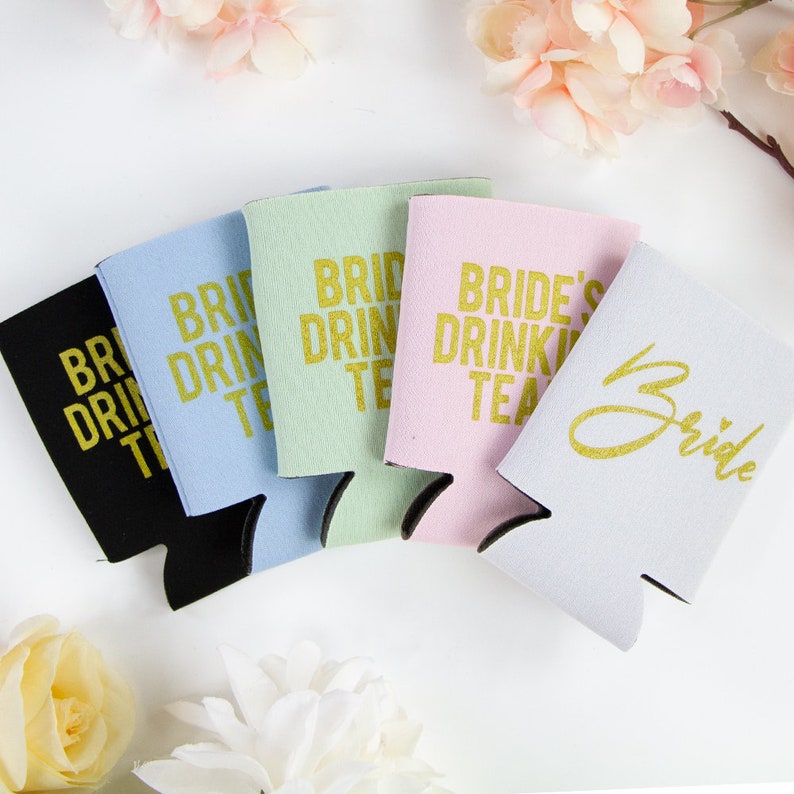 May include: Five different colored can coolers with gold lettering. The coolers say "Bride's Drinking Team", "Bride Drinking Team", "Bride Drinking Team", "Bride Drinking Team", and "Bride".