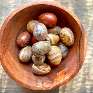 May include: A wooden bowl filled with polished, colorful, and patterned stones. The stones are various shades of brown, red, and white.