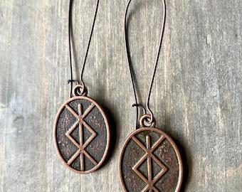 May include: A pair of copper earrings with a dark brown oval pendant featuring a stylized geometric design. The earrings have a simple wire hook closure.