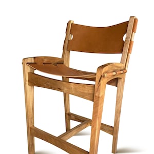 May include: A tall wooden bar stool featuring a light brown natural wood frame with visible grain. The seat and backrest are constructed from smooth, tan leather panels that are secured to the wooden structure with integrated leather straps.