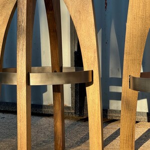 May include: A set of tall bar stools constructed from light brown wood with a visible grain. Each stool features curved legs and a circular metal footrest band with a brushed finish, providing structural support and a distinct contrast to the wood.