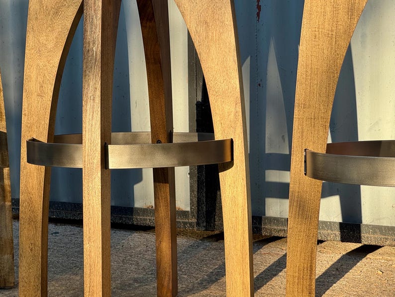 May include: A set of tall bar stools constructed from light brown wood with a visible grain. Each stool features curved legs and a circular metal footrest band with a brushed finish, providing structural support and a distinct contrast to the wood.