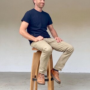 May include: A tall wooden stool with four legs and a circular seat, featuring a dark metal ring support near the base. The stool is constructed from light-toned wood with a smooth finish and a simple, functional design for elevated seating.