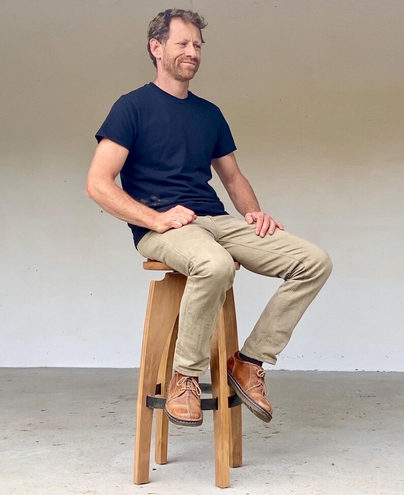 May include: A tall wooden stool with four legs and a circular seat, featuring a dark metal ring support near the base. The stool is constructed from light-toned wood with a smooth finish and a simple, functional design for elevated seating.