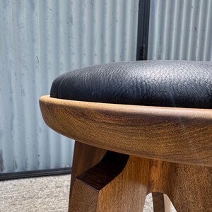 May include: A round stool featuring a dark brown wooden base with a smooth, curved frame and a black, textured leather-like upholstered seat cushion. The stool has a sturdy construction with visible wood grain patterns on the legs and frame.