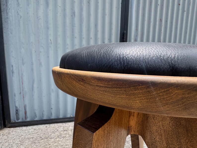 May include: A round stool featuring a dark brown wooden base with a smooth, curved frame and a black, textured leather-like upholstered seat cushion. The stool has a sturdy construction with visible wood grain patterns on the legs and frame.