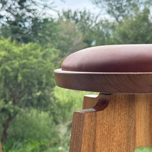May include: A wooden stool featuring a smooth, circular, reddish-brown padded seat set atop a light-toned wooden base with visible grain. The stool has a rounded frame and a sturdy construction, showcasing the natural texture of the wood.