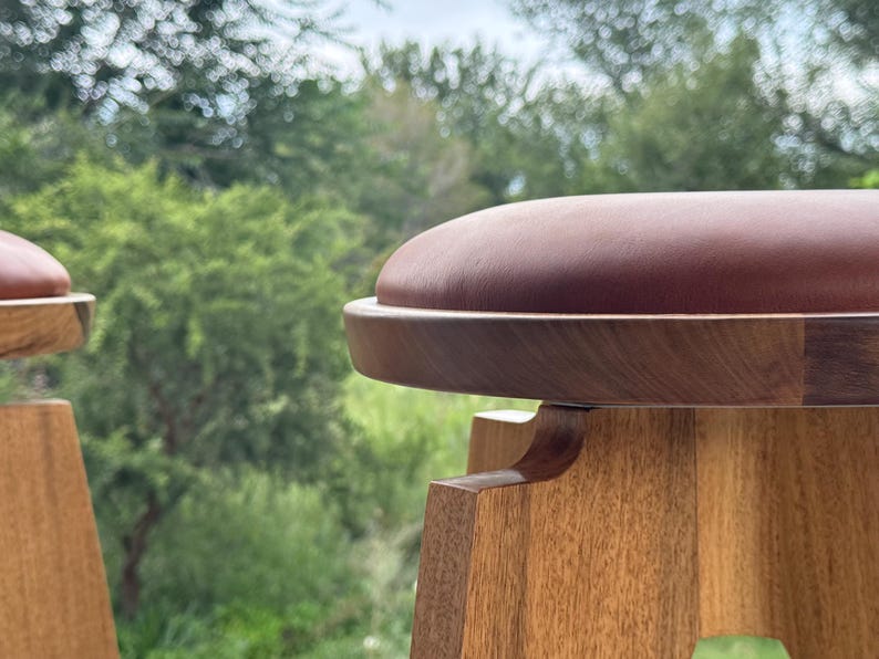 May include: A wooden stool featuring a smooth, circular, reddish-brown padded seat set atop a light-toned wooden base with visible grain. The stool has a rounded frame and a sturdy construction, showcasing the natural texture of the wood.