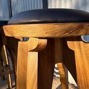 May include: A bar stool featuring a round black leather seat cushion atop a natural light brown wooden base with a thick, circular frame and sturdy, angular legs that provide a stable foundation for the elevated seating surface.