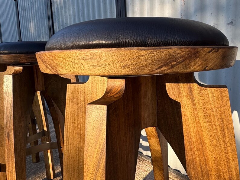 May include: A bar stool featuring a round black leather seat cushion atop a natural light brown wooden base with a thick, circular frame and sturdy, angular legs that provide a stable foundation for the elevated seating surface.