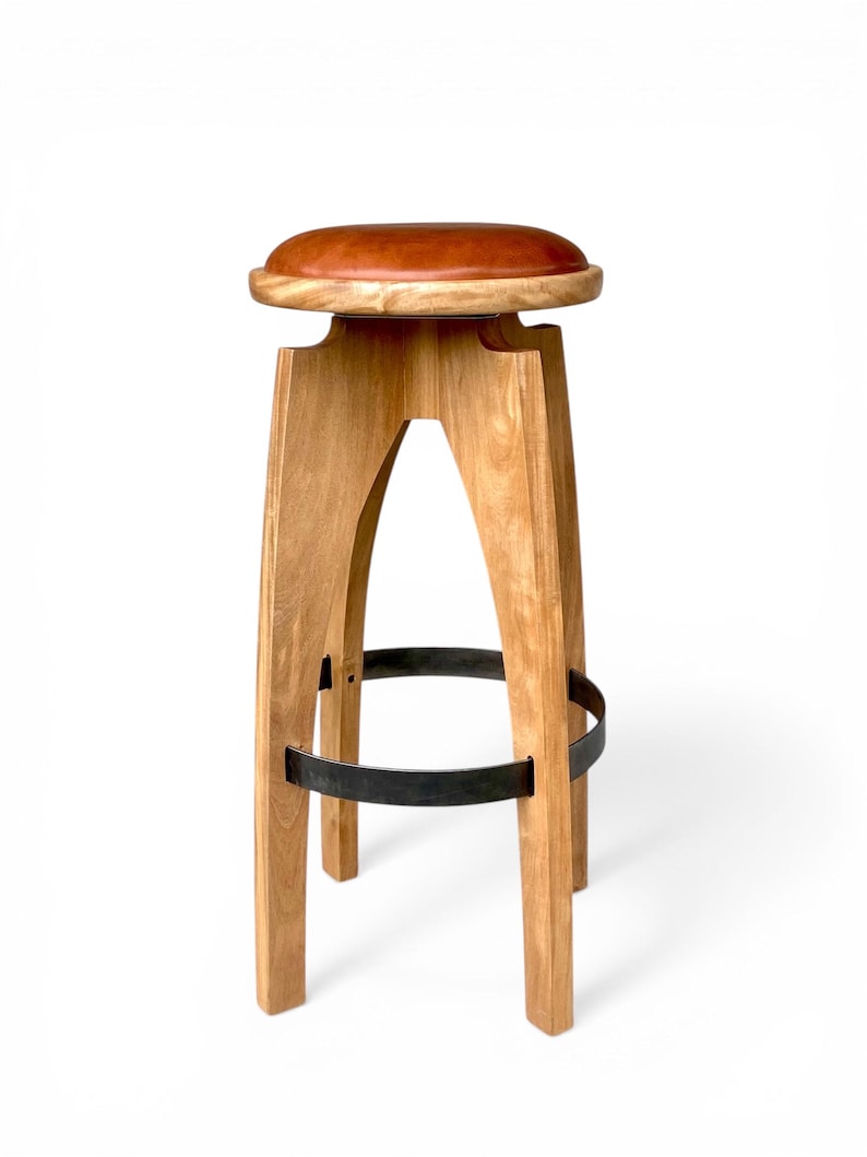 May include: A bar stool featuring a round, smooth, reddish-brown leather seat atop a light-toned wooden frame. The base consists of four curved legs connected by a circular black metal footrest ring, providing structural support and stability.
