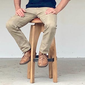 May include: A wooden stool with a round, padded seat and four angled legs connected by a circular metal footrest. The stool features a light-toned wood finish and a dark brown seat cushion, providing a stable base for sitting.