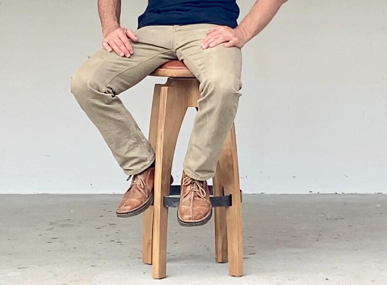 May include: A wooden stool with a round, padded seat and four angled legs connected by a circular metal footrest. The stool features a light-toned wood finish and a dark brown seat cushion, providing a stable base for sitting.
