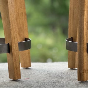 May include: Two wooden bar stools featuring light brown wood legs with a visible grain pattern. Each stool includes a curved, dark grey metal footrest ring that connects the legs, providing structural support and a distinct contrast to the wood.