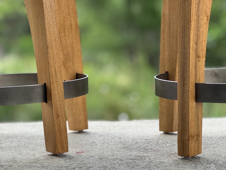 May include: Two wooden bar stools featuring light brown wood legs with a visible grain pattern. Each stool includes a curved, dark grey metal footrest ring that connects the legs, providing structural support and a distinct contrast to the wood.