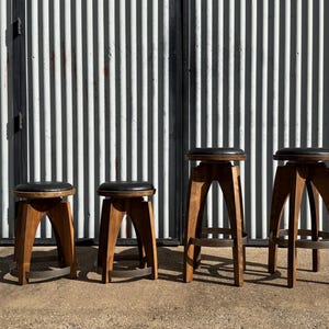 May include: Four stools with dark brown wooden legs and circular black padded seats. The two shorter stools have a lower profile, while the two taller stools feature a higher frame with a circular metal footrest ring near the base of the legs.