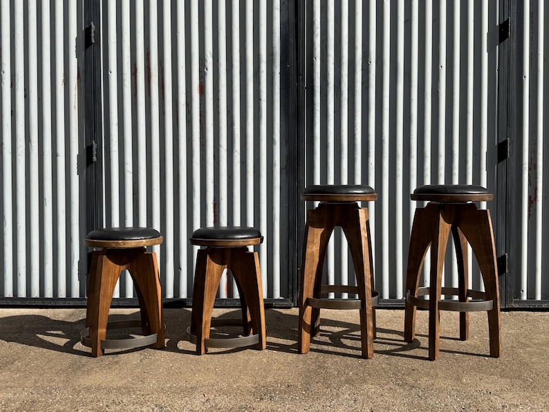 May include: Four stools with dark brown wooden legs and circular black padded seats. The two shorter stools have a lower profile, while the two taller stools feature a higher frame with a circular metal footrest ring near the base of the legs.