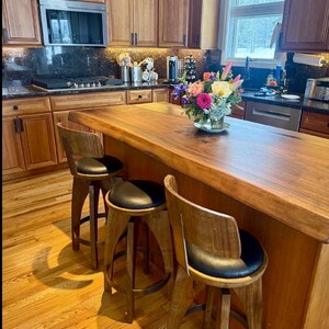 May include: Three wooden bar stools with black circular padded seats and curved backrests arranged around a large wooden kitchen island with a live edge. The island features a natural wood grain finish and a bouquet of colourful flowers in a glass vase.