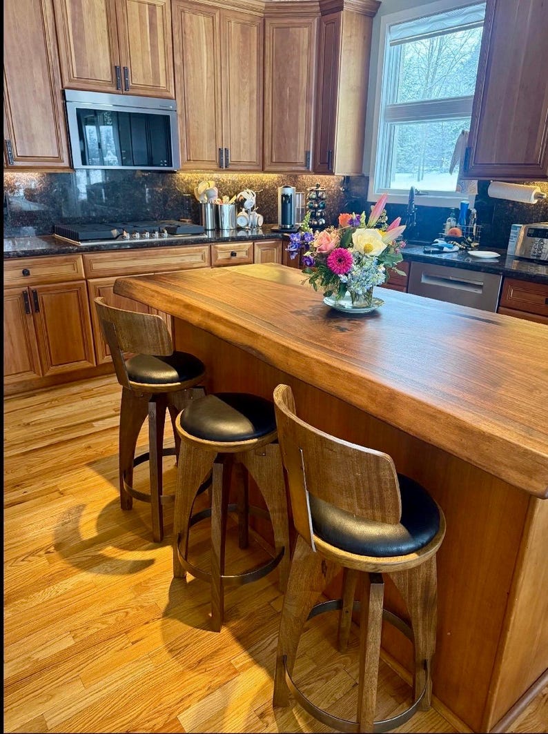 May include: Three wooden bar stools with black circular padded seats and curved backrests arranged around a large wooden kitchen island with a live edge. The island features a natural wood grain finish and a bouquet of colourful flowers in a glass vase.