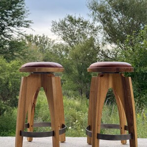 May include: Two wooden bar stools with round, dark brown leather-like seats. Each stool features four arched wooden legs connected by a circular metal footrest ring. The stools have a natural wood grain finish and a sturdy, elevated design.