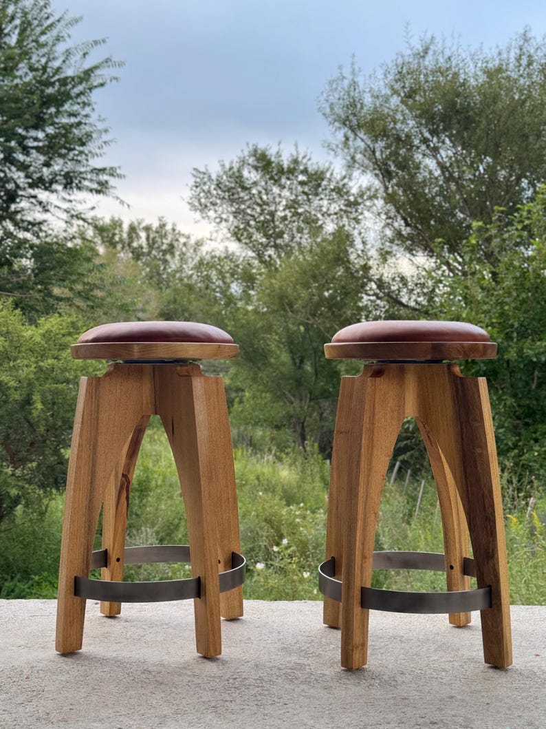 May include: Two wooden bar stools with round, dark brown leather-like seats. Each stool features four arched wooden legs connected by a circular metal footrest ring. The stools have a natural wood grain finish and a sturdy, elevated design.