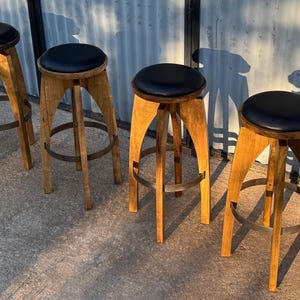 May include: Four bar stools with light brown wooden legs and circular black padded seats. Each stool features a curved leg design and a circular metal footrest ring connecting the legs near the base, providing stability and a consistent appearance.