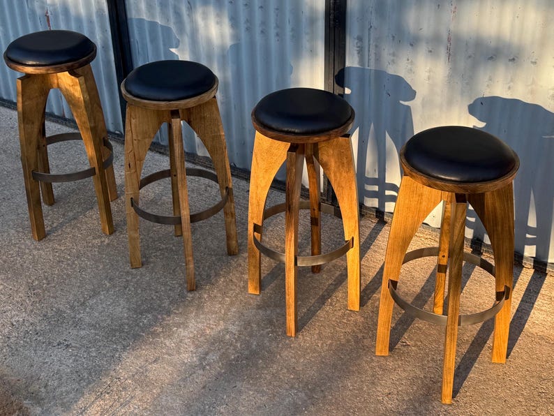 May include: Four bar stools with light brown wooden legs and circular black padded seats. Each stool features a curved leg design and a circular metal footrest ring connecting the legs near the base, providing stability and a consistent appearance.