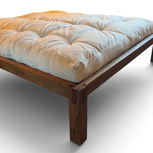 May include: A low-profile platform bed frame constructed from dark brown wood with visible joinery at the corners. The frame supports a thick, off-white tufted mattress with a grid pattern of recessed buttons across the entire surface area.