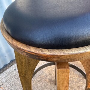 May include: A round stool featuring a black leather-like padded seat set within a circular wooden frame. The base consists of four curved wooden legs connected by a circular metal footrest, showcasing a natural wood grain finish throughout.