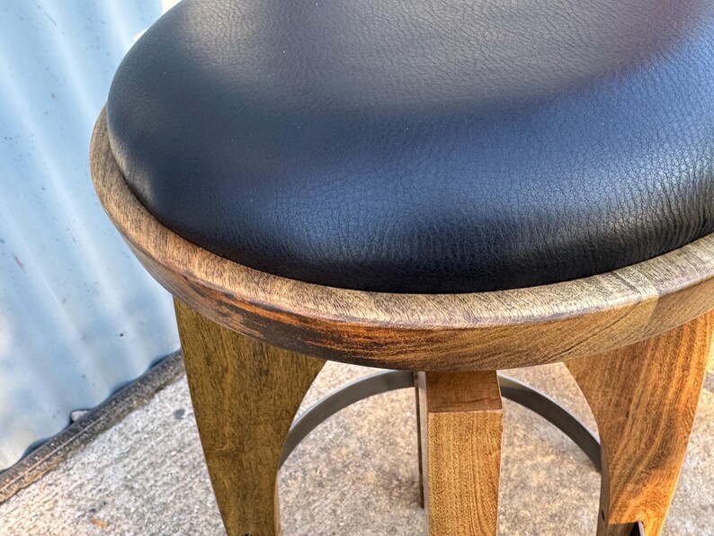 May include: A round stool featuring a black leather-like padded seat set within a circular wooden frame. The base consists of four curved wooden legs connected by a circular metal footrest, showcasing a natural wood grain finish throughout.