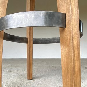 May include: A bar stool featuring four light brown wooden legs connected by a circular dark grey metal footrest. The wood grain is visible on the vertical legs, and the metal ring is securely attached to each leg, providing structural support.