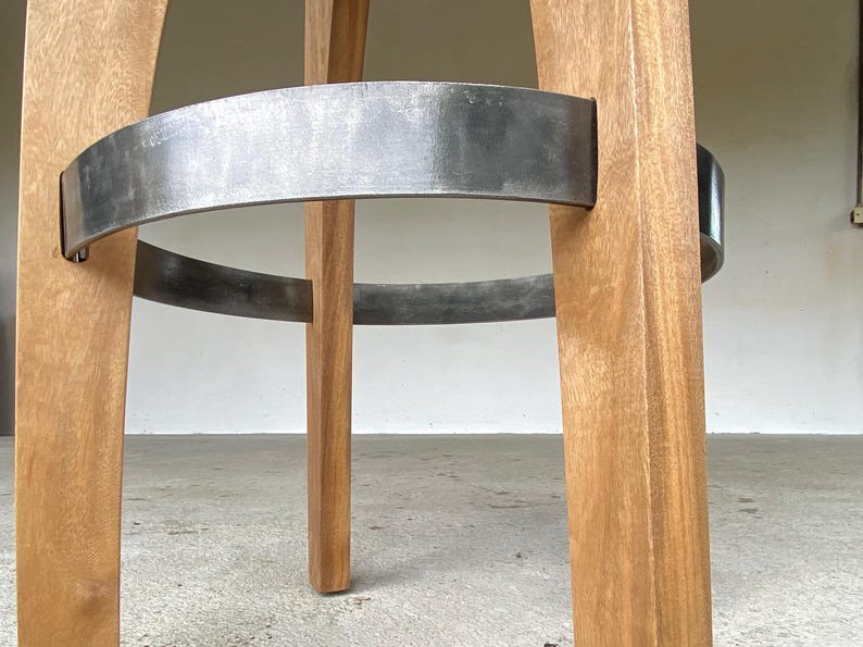 May include: A bar stool featuring four light brown wooden legs connected by a circular dark grey metal footrest. The wood grain is visible on the vertical legs, and the metal ring is securely attached to each leg, providing structural support.