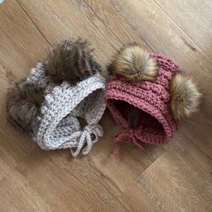 May include: Two handmade crochet hats with faux fur pom-poms. One hat is light gray with dark brown pom-poms and ear flaps. The other is dusty rose with two light brown pom-poms and ear flaps. Both hats have tie closures.