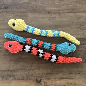 May include: Three handmade crochet snakes in yellow, blue, and red. The yellow snake has blue and green accents, the blue snake has red and green accents, and the red snake has black and white accents. Each snake has a black eye.