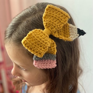 May include: A handmade crochet hair bow in the shape of a pencil. The bow is primarily mustard yellow, with a gray, pink, and cream-colored pencil tip. The pencil eraser is black. The bow is attached to a hair clip.