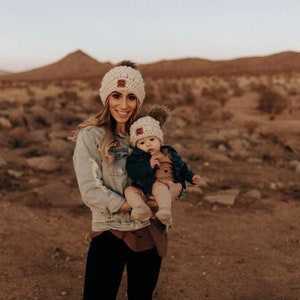 May include: Cream-colored knit beanie hats with a brown pom-pom and a small patch that says "MA MA". One hat is worn by an adult, the other by a baby. The adult is wearing a denim jacket and black pants.